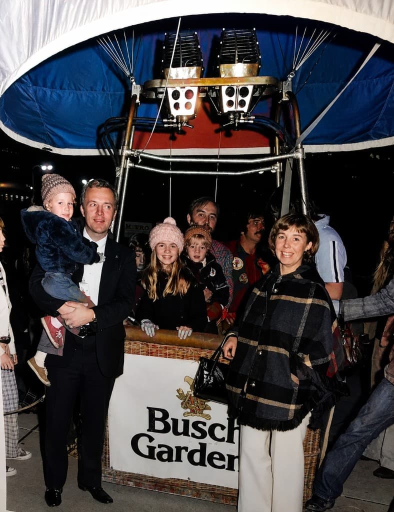 A man in a tuxedo holding a child beside a hot-air balloon basket draped with a Busch Gardens banner, surrounded by children and adults at an evening event.