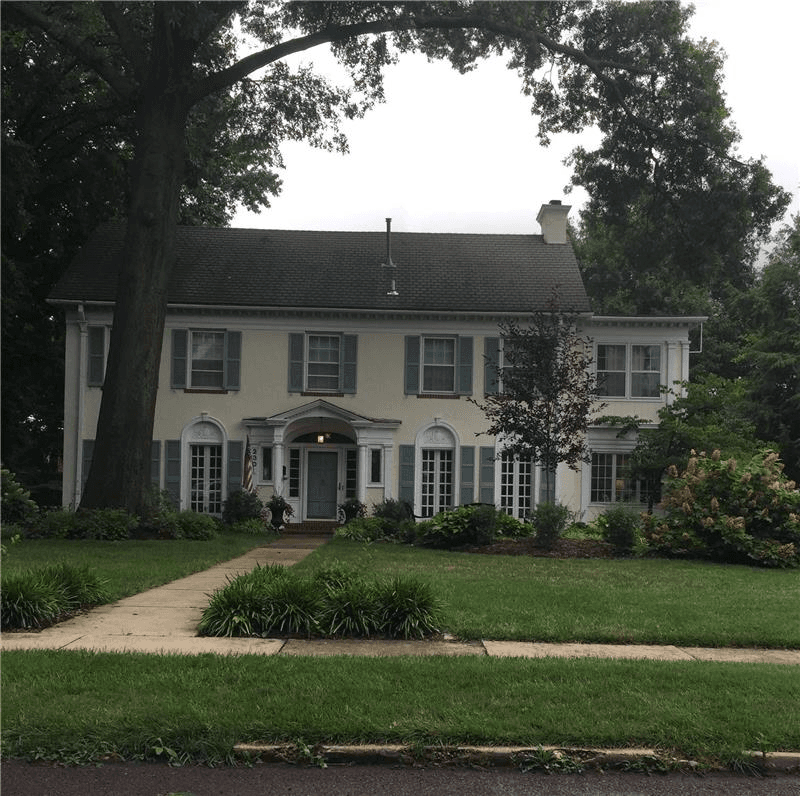 Two-story house with pale stucco walls, green shutters, arched door surrounds, and a large shade tree in the front yard.