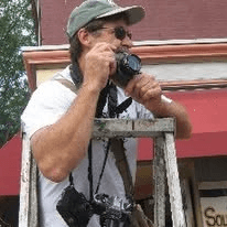 A man standing on a ladder holding a camera, with a second camera around his neck.
