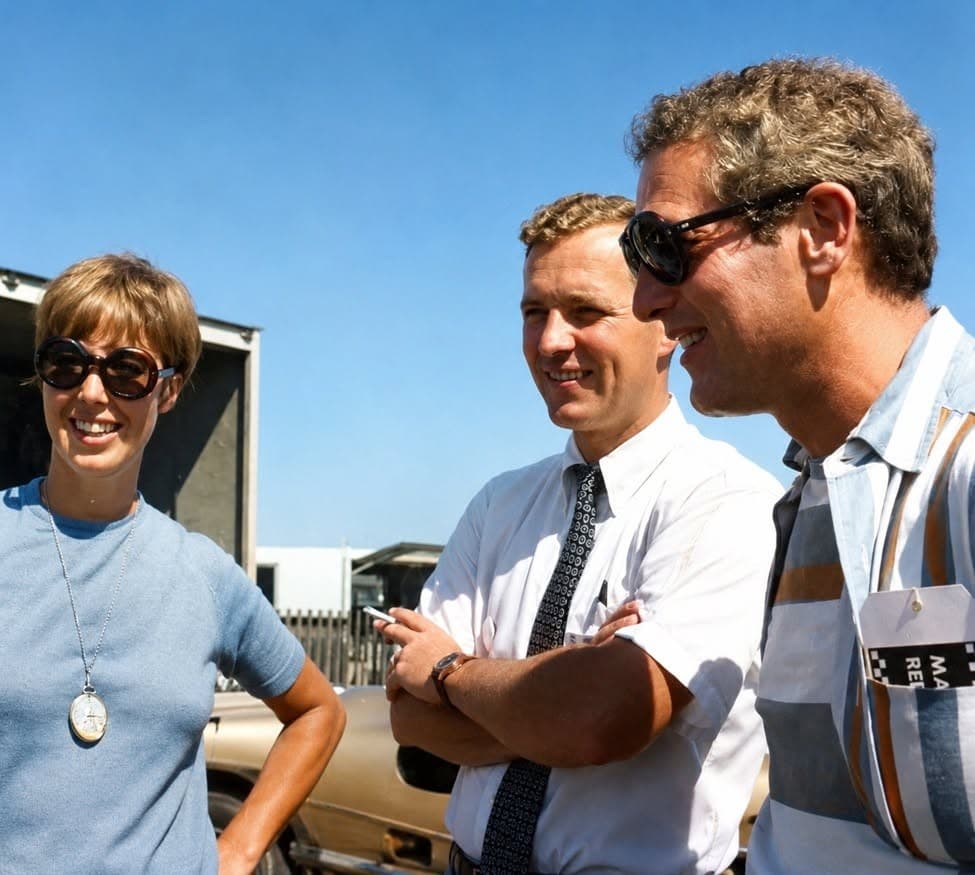 Vintage color photograph at a racetrack: a smiling woman in round sunglasses, a man in a white shirt and tie with folded arms, and a man in sunglasses wearing a media credential, beside a race car and transporter under a blue sky.